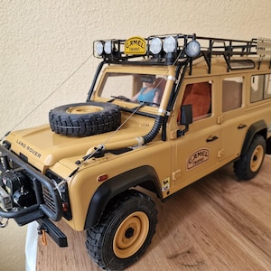 May include: A tan Land Rover Defender model with a black roof rack, spare tyre, and "Camel Trophy" branding. The model has black bumpers, a winch, and spotlights. The text "Land Rover" is visible on the bonnet.