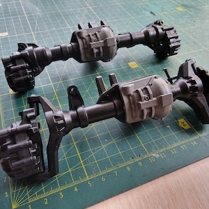 以下が含まれることがあります： 緑色のカッティングマットの上に、黒とグレーの模型車の車軸が2つあります。 各車軸には、中央のチューブ、ギアハウジング、詳細なコンポーネントがあり、趣味や模型製作をテーマにしています。