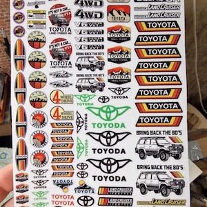 Może przedstawiać: Arkusz naklejek z różnymi wzorami i logo Toyota Land Cruiser. Naklejki zawierają tekst, taki jak "4WD", "V8 Turbo" i "Bring Back the 80's". Schemat kolorów obejmuje kolory czerwony, pomarańczowy, żółty, czarny i biały.