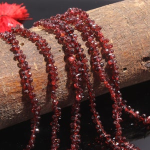 May include: Multiple strands of deep red, faceted garnet beads are draped over a textured, cylindrical wooden log. The small, translucent gemstone beads catch the light, displaying a dark crimson hue and a polished, multi-faceted surface texture.