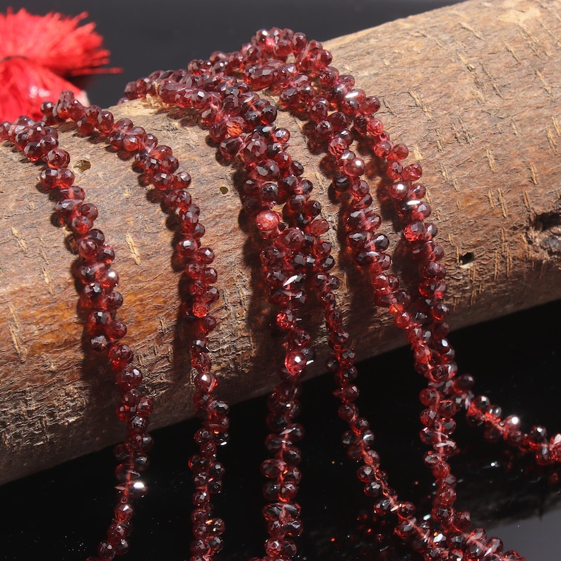 May include: Multiple strands of deep red, faceted garnet beads are draped over a textured, cylindrical wooden log. The small, translucent gemstone beads catch the light, displaying a dark crimson hue and a polished, multi-faceted surface texture.