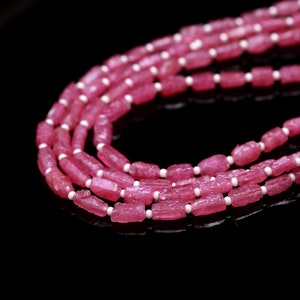 Pode incluir: Um colar de várias camadas com contas cilíndricas de pedra preciosa rosa bruta, intercaladas com pequenas contas esféricas brancas e lisas. O colar está disposto numa formação curva e sobreposta, exibindo a textura das pedras rosas.