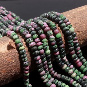 Könnte beinhalten: Mehrere Stränge aus runden, scheibenförmigen Perlen mit einem gesprenkelten Muster in tiefem Magenta, Waldgrün und Schwarz. Die Perlen sind auf langen, durchgehenden Schnüren aufgereiht und weisen eine glatte, polierte Textur sowie eine abwechslungsreiche Farbverteilung auf.