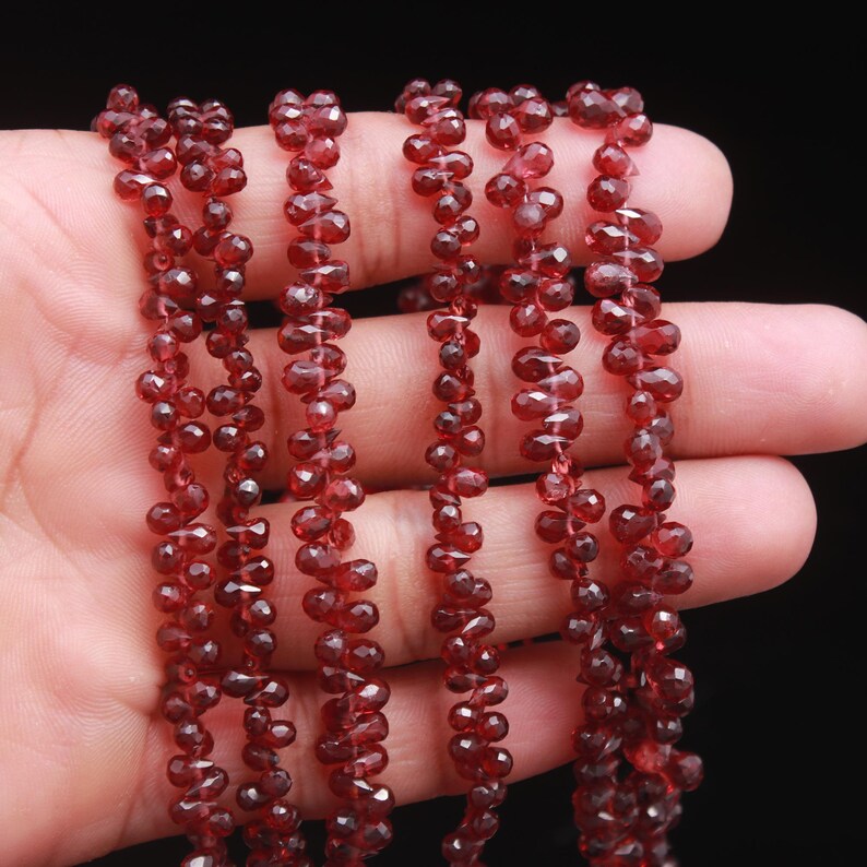 May include: Multiple strands of deep red, faceted garnet beads shaped like small teardrops. Each bead features a polished, reflective surface that catches the light, arranged in vertical lines on thin, clear stringing material.