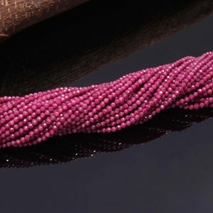 May include: A long strand of small, round, faceted beads in a deep magenta-pink color. The beads are uniform in size and shape, featuring a reflective, multi-faceted surface that catches the light, arranged in a dense, continuous horizontal line.