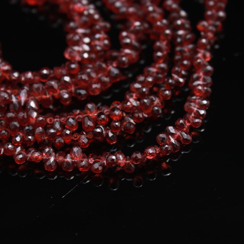 May include: A strand of deep red, faceted, teardrop-shaped beads arranged in multiple overlapping loops. The translucent, gemstone-like beads feature a polished surface that reflects light, creating a vibrant, sparkling appearance against a dark background.