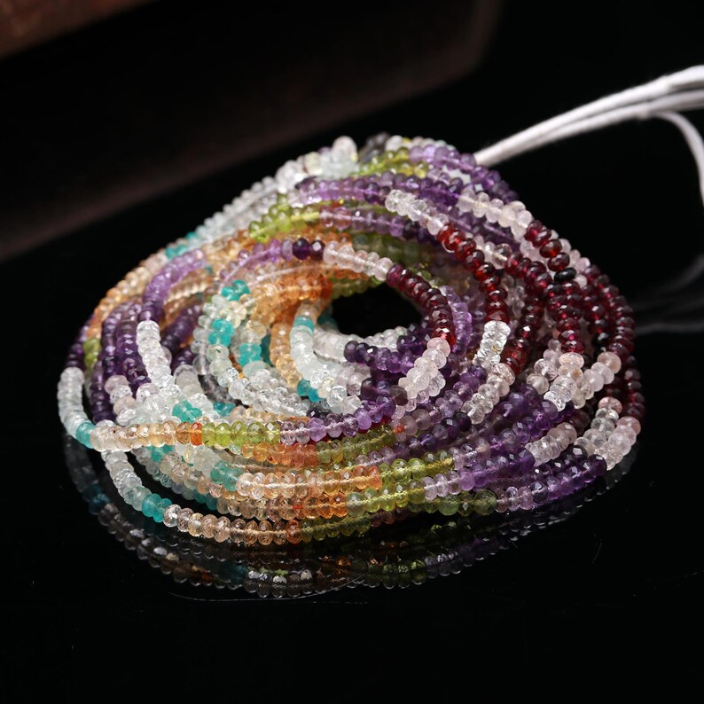 May include: A coiled strand of small, faceted gemstone beads in a variety of colors including deep red, purple, light green, blue, and clear white. The beads are strung on a thin white cord, creating a circular arrangement with a central opening.