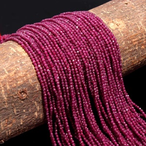 Peut inclure: Un long collier composé de petites perles facettées de couleur magenta profond, disposées en plusieurs rangées parallèles. Les perles ont une surface texturée et réfléchissante, drapées sur un support cylindrique en bois brun texturé.