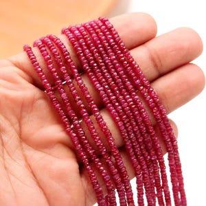 May include: Multiple strands of small, faceted, deep pinkish-red ruby beads arranged in parallel lines. Each bead features a circular, flattened shape with a reflective, textured surface, creating a consistent, uniform appearance across all the strands.