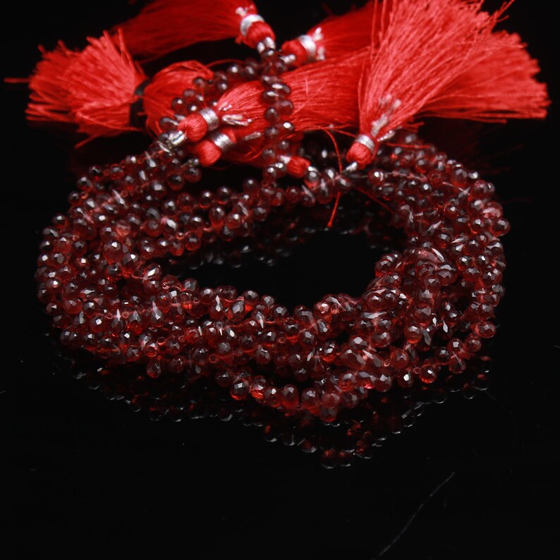 May include: A strand of deep red, faceted, teardrop-shaped gemstone beads arranged in a circular loop. The beads are strung together and accented with multiple bright red decorative tassels featuring silver-toned metal caps at the top.