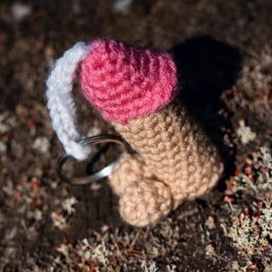 May include: A handmade, crocheted keychain shaped like a penis. The top is pink, the shaft is tan, and the base is a lighter tan. A white loop extends from the top, and a silver keyring is attached. The keychain is set against a natural, textured background.