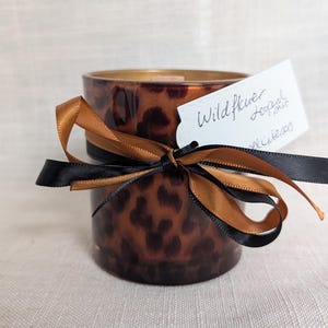 May include: A cylindrical candle holder with a leopard print pattern. It is decorated with a black and gold ribbon and a white tag that reads "Wildflower Leopard Print". The candle holder sits on a neutral background.