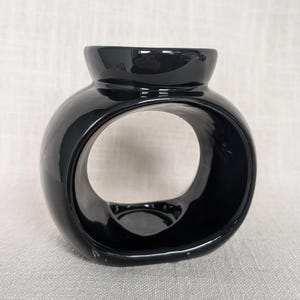 May include: A black ceramic oil diffuser with a distinctive circular design. The diffuser has a rounded body with a large central opening and a small opening at the top. The surface is smooth and reflective.