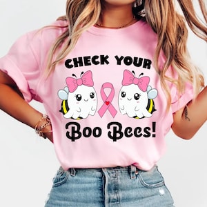 May include: Pale pink t-shirt featuring the slogan "CHECK YOUR Boo Bees!" and a graphic of two cartoon bees with pink bows and a pink ribbon. The bees have white bodies, black eyes, and yellow and black wings.