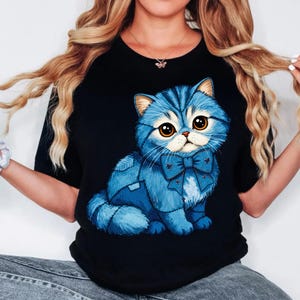 Könnte beinhalten: Schwarzes T-Shirt mit einer Cartoon-Illustration einer blauen Katze mit Fliege. Die Katze hat große Augen und eine flauschige Textur. Das Design ist zentral platziert und bietet eine skurrile und ansprechende Grafik.