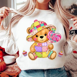 May include: White sweatshirt featuring a cartoon teddy bear wearing a patchwork hat and outfit, holding a heart-shaped lollipop. Two cupcakes are on the bottom of the sweatshirt. The bear has a cheerful expression. The design is colorful and whimsical.