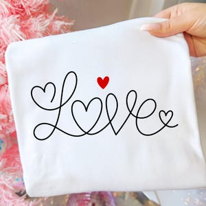 Op de afbeelding: Een wit stoffen item met het woord "Love" in een zwart, cursief lettertype. De letter "O" is vervangen door een rood hart en het woord is versierd met hartvormen.