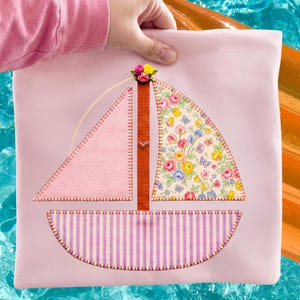 Op de afbeelding: Een roze stoffen zeilbootontwerp met een bloemenzeil en gestreepte basis. De boot heeft een bruine leren mast met een klein hartje en is bekroond met een tros kleurrijke bloemen. Het ontwerp is op een lichtroze achtergrond gestikt.