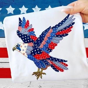 May include: White fabric featuring a patriotic eagle design. The eagle's wings are spread, showcasing a patchwork of red polka dots, blue gingham, and navy blue polka dots. The eagle's head has blue and white accents with star details.