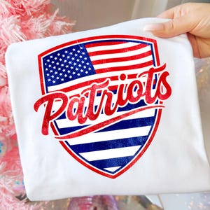 May include: White t-shirt featuring a shield design with the American flag and the word "Patriots" in red script. The design incorporates red, white, and blue colors, evoking a patriotic theme. The shirt is held up by a hand.