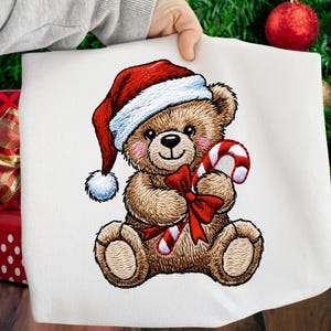 Peut inclure: Tissu blanc brodé avec un ours en peluche portant un bonnet de Père Noël et tenant un sucre d'orge. L'ours a un nœud rouge et des joues roses. Le motif utilise du fil marron, rouge, blanc et bleu, parfait pour les décorations de Noël.