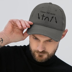 Puede incluir: Gorra de béisbol gris desgastada con el texto "I know THE MAN!" y símbolos religiosos. La gorra tiene un aspecto desgastado con una visera curva y una correa ajustable. Un accesorio casual.