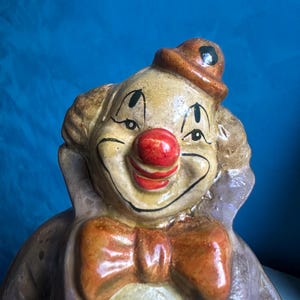 May include: A vintage clown figurine with a smiling face, red nose, and orange bow tie. The clown wears a small brown hat and a lavender coat. The figurine has a painted, aged appearance.
