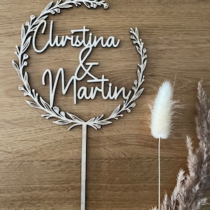 May include: A wooden cake topper with a floral wreath design. The topper features the names "Christina & Martin" in a cursive font.