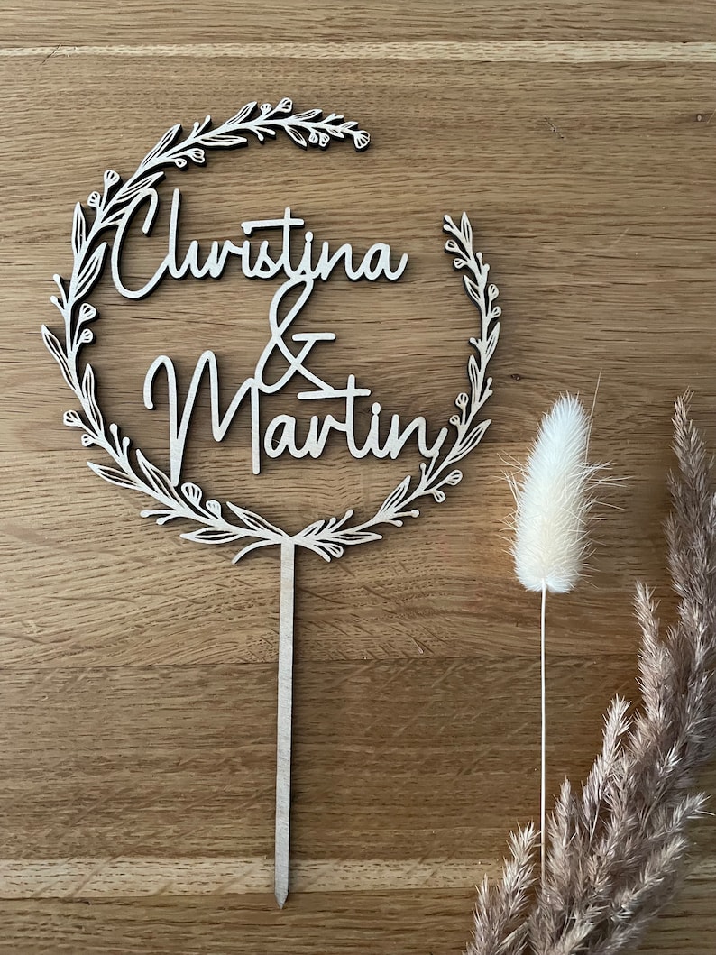 May include: A wooden cake topper with a floral wreath design. The topper features the names "Christina & Martin" in a cursive font.