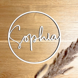 Könnte beinhalten: Ein weißer Holz-Kreis mit dem Namen "Sophia" in kursivschrift. Der Kreis befindet sich auf einer hellbraunen Holzoberfläche.