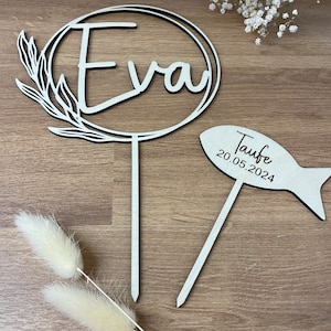 Op de afbeelding: Houten taarttopper met een cirkel en de naam "Eva" erin. Een tweede topper is in de vorm van een vis met de tekst "Taufe 20.05.2024".
