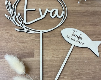 Op de afbeelding: Houten taarttopper met een cirkel en de naam "Eva" erin. Een tweede topper is in de vorm van een vis met de tekst "Taufe 20.05.2024".