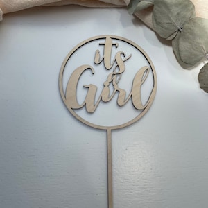Może przedstawiać: Drewniany topper na tort z okrągłym wzorem i napisem "it's a girl" w ozdobnym stylu pisma.