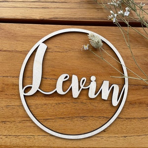 Könnte beinhalten: Ein Holz-Kreis mit dem Namen "Levin" ausgeschnitten in einer kursiven Schrift. Der Kreis liegt auf einer Holzoberfläche.