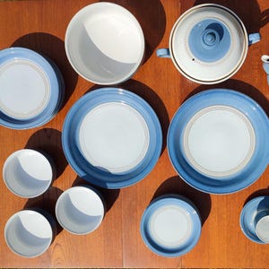 May include: A vintage blue and white ceramic tableware set, including plates, bowls, cups, saucers, a teapot, and a serving dish. The set has a light blue border with a white centre and a decorative trim.