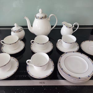 May include: A white porcelain tea set with blue and gold detailing. The set includes a teapot, sugar bowl, creamer, cups, saucers, and plates. The tea set is displayed on a black surface.
