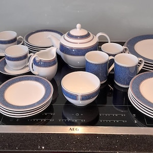 May include: A full dinner service in white and blue. The set includes cups, saucers, plates, bowls, a teapot, and a milk jug. The plates and cups have a blue patterned border. The set is arranged on a black surface.