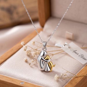 Puede incluir: Un collar de tono plateado con un colgante de una figura estilizada abrazando a un perro de tono dorado. El colgante cuelga de una cadena fina. Una pequeña etiqueta blanca unida a la joya muestra el texto Amor Eterno en letra cursiva.