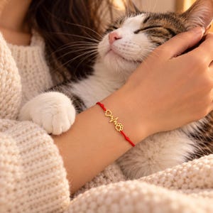 May include: A delicate red string bracelet featuring a gold-toned metal charm that displays a heart, an electrocardiogram heartbeat line, and a paw print. The bracelet is worn on a wrist, positioned near a white and brown cat.