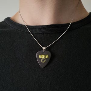 May include: A silver-toned chain necklace featuring a black guitar pick pendant. The pendant is printed with the yellow text NIRVANA and a yellow smiley face graphic. The necklace is worn against a plain black shirt, showcasing the pendant design.