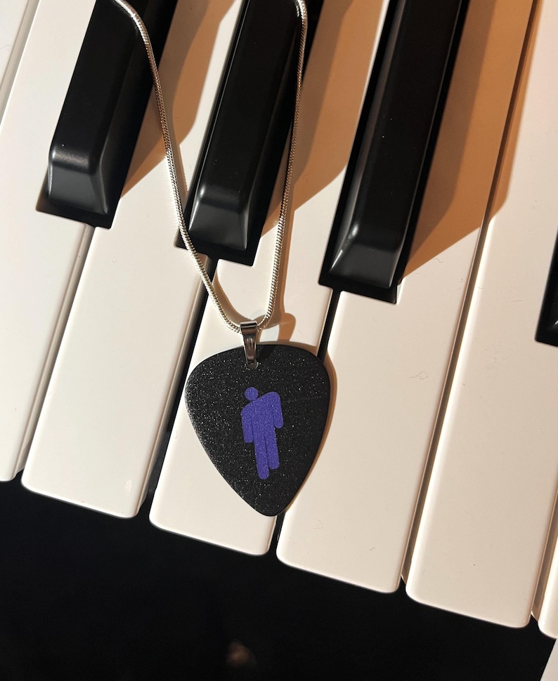 May include: A black guitar pick necklace with a purple silhouette of a person. The necklace is on a white and black keyboard.