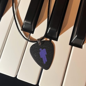 May include: A black guitar pick necklace with a purple silhouette of a person. The necklace is lying on a white and black keyboard.