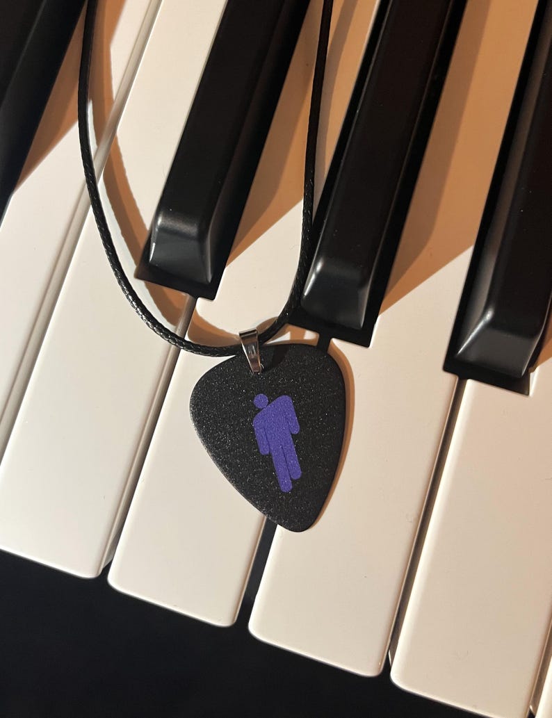 May include: A black guitar pick necklace with a purple silhouette of a person. The necklace is lying on a white and black keyboard.