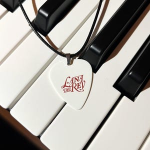 Peut inclure: Un collier avec un m&eacute;diator de guitare blanc et un logo rouge "Lana Del Rey". Le collier est sur un cordon noir et repose sur un clavier blanc et noir.