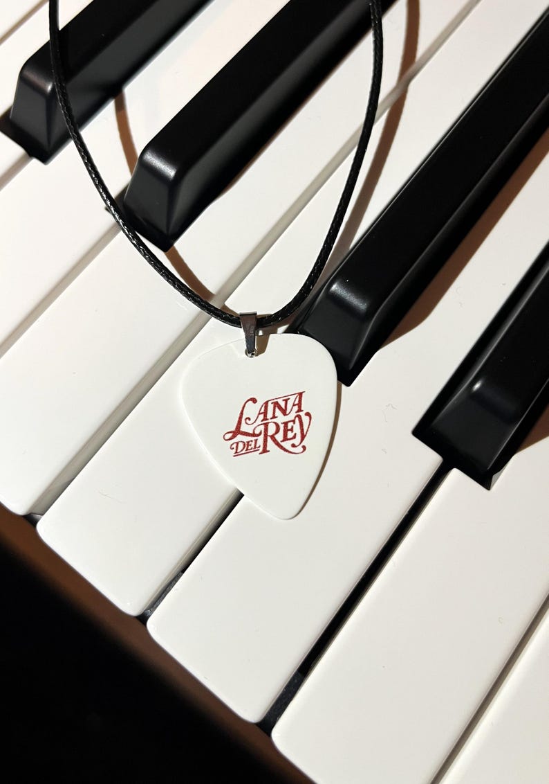 Peut inclure: Un collier avec un m&eacute;diator de guitare blanc et un logo rouge "Lana Del Rey". Le collier est sur un cordon noir et repose sur un clavier blanc et noir.