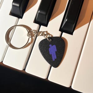 May include: A black guitar pick keychain with a purple silhouette of a person. The keychain is attached to a silver key ring and is lying on a white keyboard.