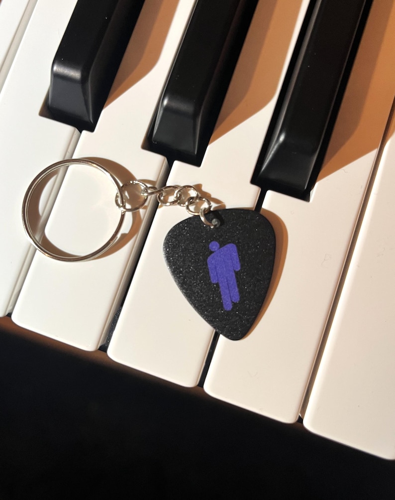 May include: A black guitar pick keychain with a purple silhouette of a person. The keychain is attached to a silver key ring and is lying on a white keyboard.