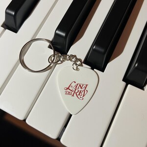Peut inclure: Porte-cl&eacute;s en forme de m&eacute;diator de guitare blanc avec un logo "Lana Del Rey" rouge sur un clavier de piano blanc.