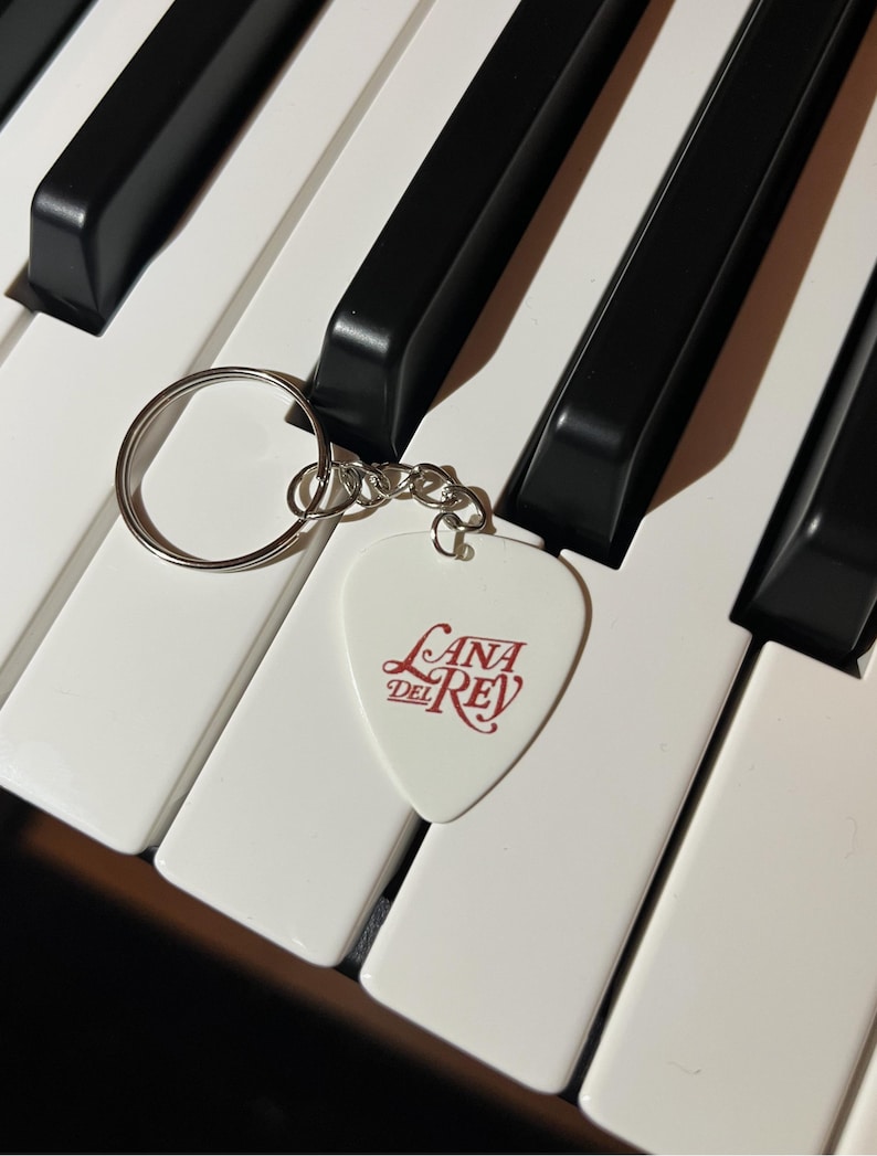 Peut inclure: Porte-cl&eacute;s en forme de m&eacute;diator de guitare blanc avec un logo "Lana Del Rey" rouge sur un clavier de piano blanc.