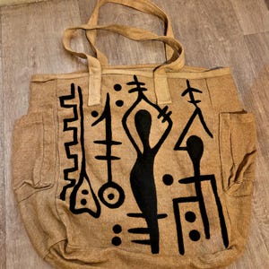 Peut inclure: Un sac fourre-tout beige doté de deux bandoulières et de poches latérales. L'avant du sac est orné d'un grand motif graphique peint à la main en noir, composé de symboles abstraits, de formes géométriques et d'une silhouette humaine stylisée.